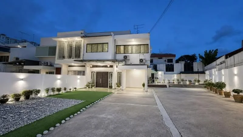 Exterior type: detached bungalow; features: white painted facade, flat roof, 2-storey building, manicured lawn, landscaped garden with stones, outdoor lighting, multiple air conditioning units visible, paved driveway, fenced perimeter, decorative potted plants; style: modern.