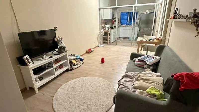 Living features: grey fabric sofa, white TV console, wall-mounted TV, area rug, wooden flooring, ceiling fan, decorative shelf, toys scattered on the floor, laundry on the sofa; style: modern. Kitchen features: open kitchen layout, glass sliding doors, kitchen cabinets, kitchen countertop; style: modern.