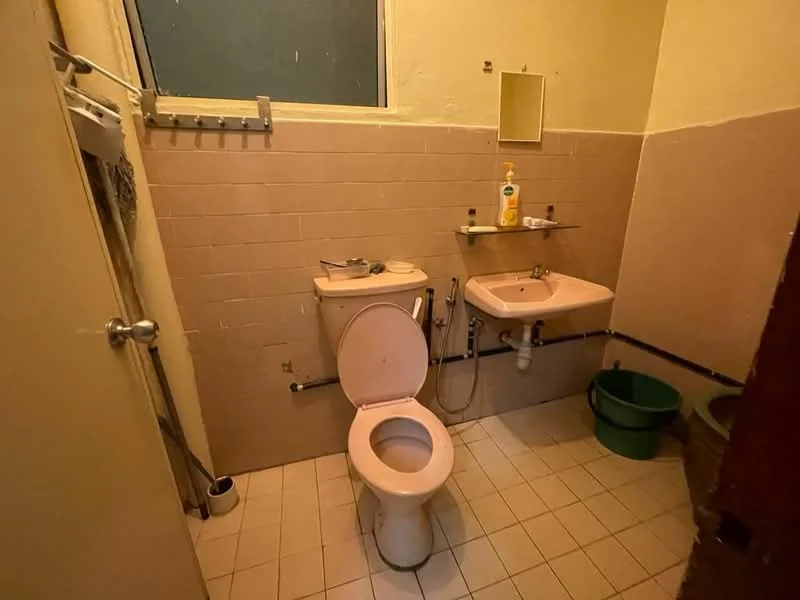 Bathroom features: pink toilet, pink sink, mirror above sink, wall-mounted soap dispenser, tiled walls in light beige, tiled flooring in white, shower hose attached to sink, window for natural light, metal towel rack, green bucket.