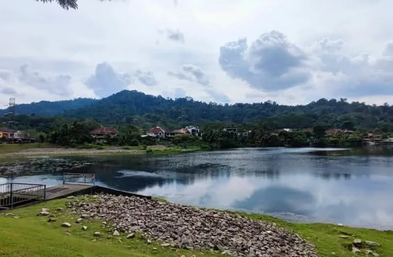 Exterior type: landscape garden; features: lake view, manicured lawn, rocky shoreline, surrounding hills, residential houses in background, cloudy sky; style: tropical-modern.