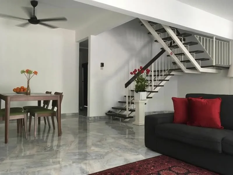 Living features: grey fabric sofa, red decorative cushions, area rug with geometric pattern, marble flooring, ceiling fan, wall-mounted light switch, decorative vase with flowers, coffee table; style: modern. Dining features: round wooden dining table, 4 green upholstered dining chairs, fruit bowl with oranges, white walls, natural light from windows; style: modern.