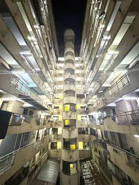 Exterior type: mid-rise condominium; features: concrete facade, multiple balconies, central service column, illuminated corridors, metal railings, exposed concrete surfaces, windows with yellow lighting, interior courtyard, multiple floors, nighttime lighting; style: modern.