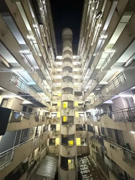 Exterior type: mid-rise condominium; features: concrete facade, exposed concrete columns, balconies with metal railings, central ventilation shaft, multiple floors visible, night lighting, windows with yellow light, staircase access, interior courtyard view; style: modern.