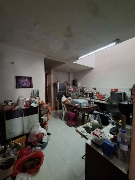 Room features: white painted walls, tile flooring, ceiling with visible stains, overhead fluorescent lighting, wooden dining table, plastic chairs, refrigerator, fan, various kitchen appliances, cluttered with bags and containers, open layout, kitchen cabinetry, multiple visible items on surfaces.