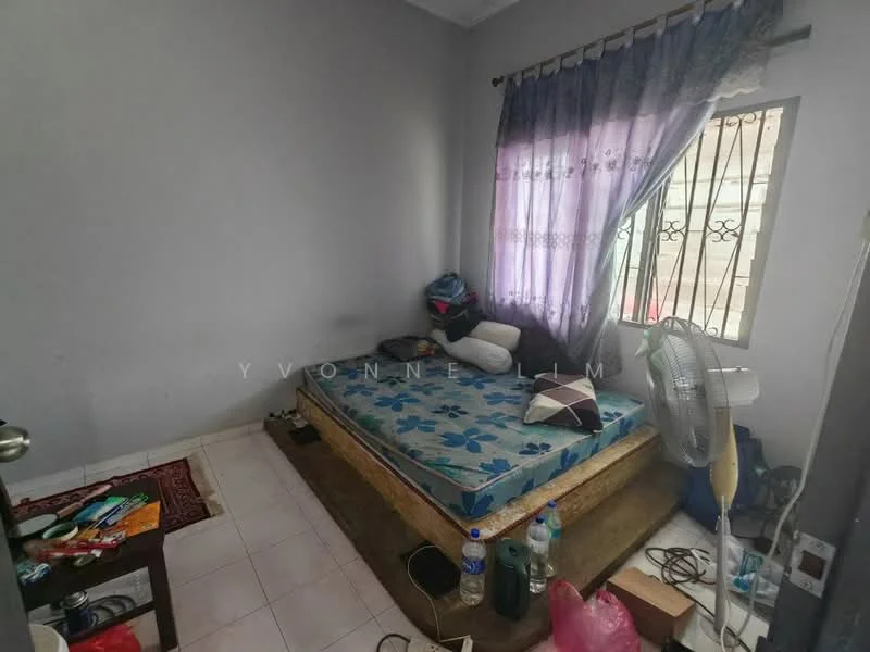 Bedroom features: single bed with floral bedding, wooden bed frame, white painted walls, tiled flooring, curtains in purple with lace trim, window with security bars, floor fan, small wooden table, various personal items on the floor, bottled water, decorative pillows.
