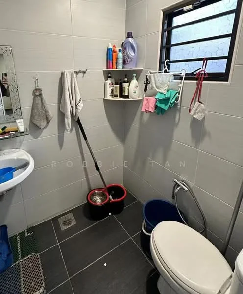 Bathroom features: grey tiled walls, black tiled flooring, white sink with chrome faucet, wall-mounted mirror, toilet, bidet spray, shelf with toiletries, hanging towel rack, window for natural light, cleaning supplies in blue bucket, pet bowls, hand towel, shower area with drain; style: modern.