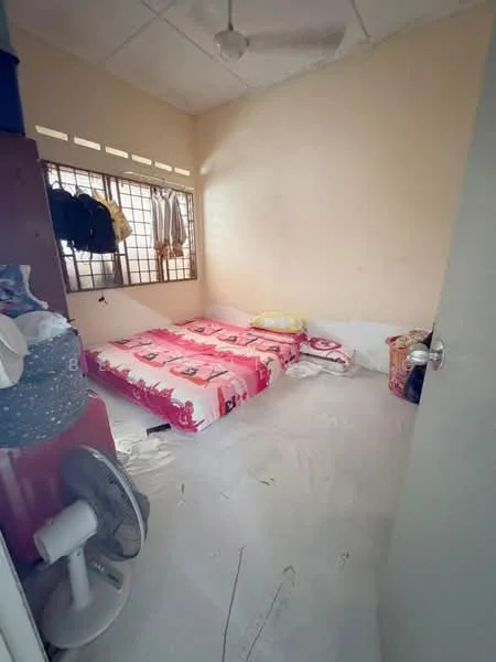 Bedroom features: single bed with pink floral bedding, white walls, ceiling fan, window with grid pattern, clothes hanging on hooks, wooden wardrobe, floor covered with white plastic sheet, small fan on the floor.
