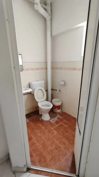 Bathroom features: white toilet, single-sink vanity with mirror, tile flooring in brown, white walls, window for natural light, toilet paper holder, plumbing pipe exposed, waste bin.
