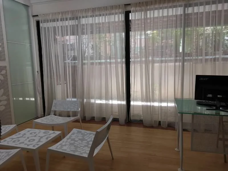 Room features: large floor-to-ceiling windows, sheer curtains, wooden flooring, glass-top desk, white chairs with floral pattern, minimalist decor, natural light, indoor plants visible outside; style: modern; view: garden.