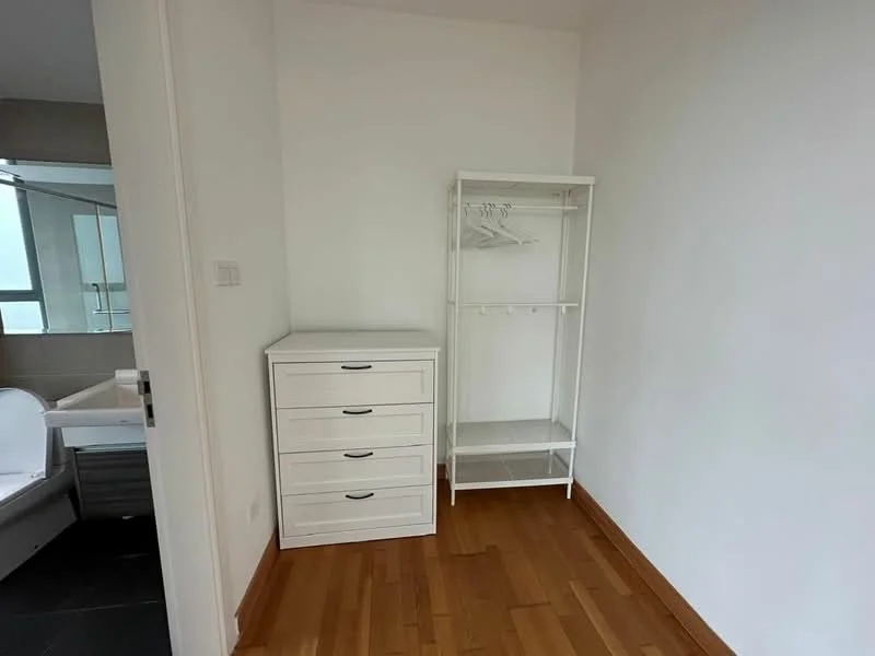 Room features: white painted walls, wooden flooring, 4-drawer white dresser, open clothing rack, natural light from window, minimalist decor; style: modern.