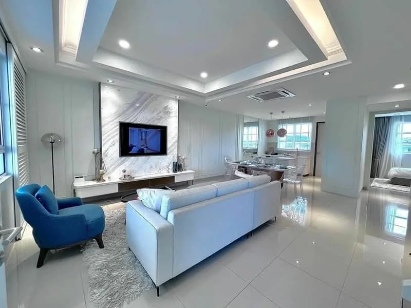 Living features: white painted walls, tray ceiling with recessed lighting, marble feature wall, large flat-screen TV, 3-seater white fabric sofa, blue accent chair, area rug, glass coffee table, decorative vases, LED ceiling lights, sliding glass door, window with sheer curtains, dining area visible in background; style: modern.