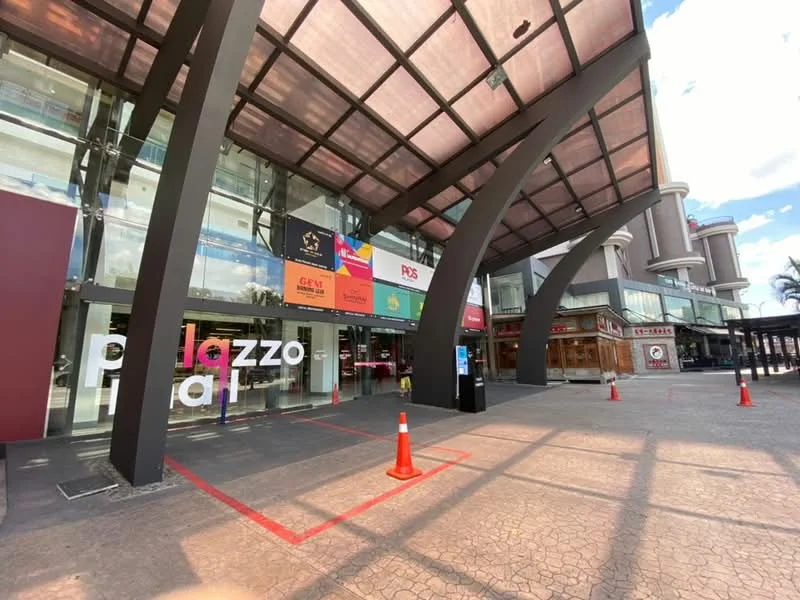 Exterior type: commercial building; features: glass facade, metal awning, large entrance, red and white signage, paved walkway, traffic cones, outdoor seating area, surrounding palm trees, multiple storeys visible, modern architectural design; style: modern.