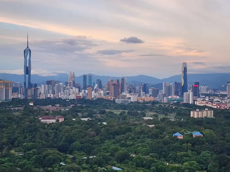 Exterior features: city skyline, greenery in foreground, mountain backdrop, various high-rise buildings, Petronas Twin Towers, Merdeka 118, urban landscape, cloudy sky; style: modern.