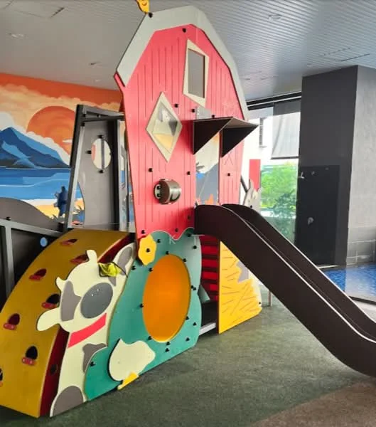 Exterior type: children's playground; features: colorful play structure, slide, climbing elements, animal-themed decorations, safety flooring, surrounding greenery, wall mural with mountains and sunset; style: modern.