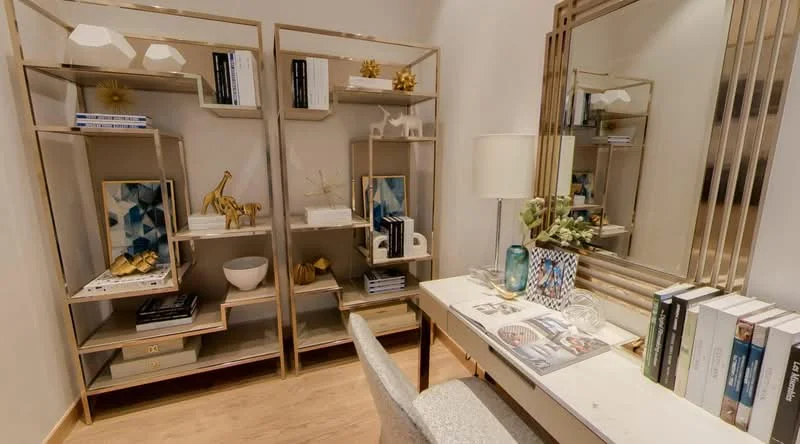 Study features: gold metal shelving units, decorative items on shelves, white ceramic bowl, framed artwork, desk with white surface, silver table lamp, mirror with gold frame, wooden flooring, decorative books, glass decorative item; style: modern.
