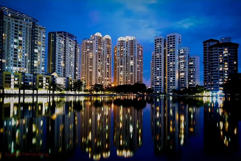 Exterior type: high-rise condominium; features: glass curtain wall, illuminated facade, surrounded by water, reflections on water surface, multiple high-rise buildings, nighttime lighting, landscaped grounds, urban skyline, mature trees in foreground; style: modern.
