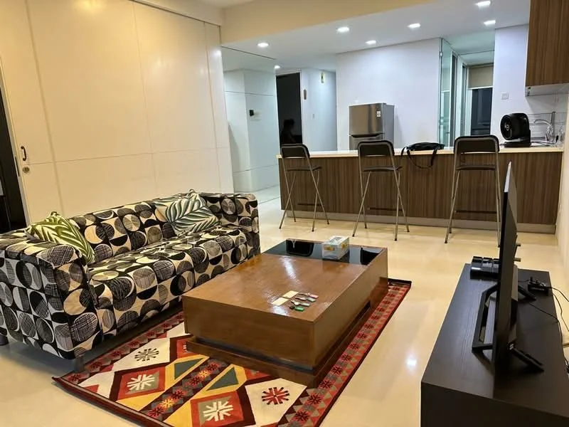 Living features: 3-seater fabric sofa with geometric pattern, wood coffee table, area rug with ethnic pattern, wall-mounted TV, bar stools at kitchen counter, recessed downlights, cream tiled flooring, open layout to kitchen, kitchen island, fridge visible in background; style: modern.