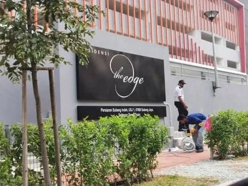 Exterior features: signage with the name 'the edge', grey painted facade, landscaped greenery with shrubs, paved walkway, outdoor lighting, visible construction or maintenance activity; style: modern.