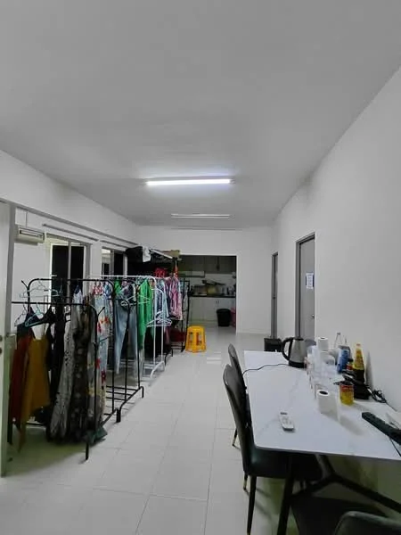 Room features: white painted walls, tile flooring, fluorescent ceiling lights, clothes rack with hanging garments, table with chairs, kitchen area visible in the background, open layout, utility items on the table.