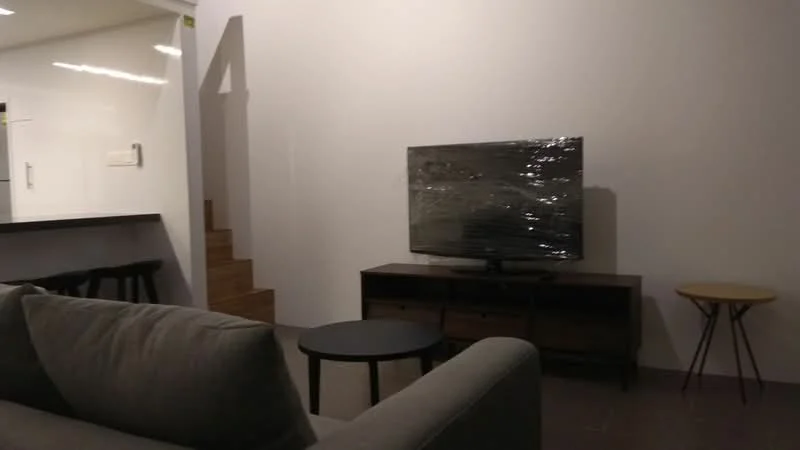 Living features: grey fabric sofa, wooden TV console, wall-mounted TV wrapped in plastic, black round coffee table, light wood side table, white painted walls, tile flooring, staircase in background; style: modern.