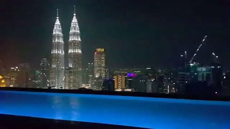 Exterior type: swimming pool deck; features: infinity pool with illuminated edge, city skyline view, night lighting, surrounding high-rise buildings, Petronas Twin Towers visible, urban landscape, decorative lighting around pool area; style: modern.