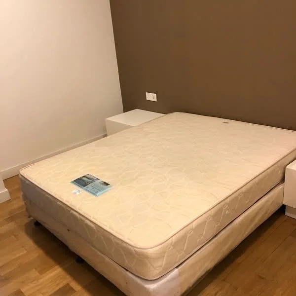 Bedroom features: queen-size bed base, mattress without bedding, light brown accent wall, white painted walls, wooden flooring, two white bedside tables, one electrical outlet on the wall; style: modern.