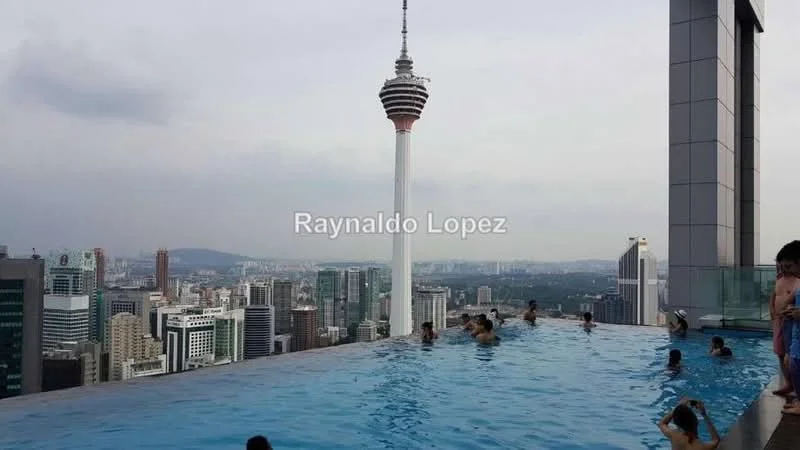 Exterior type: high-rise condominium; features: infinity pool with city skyline view, pool deck with loungers, surrounded by urban landscape, KL Tower visible in background, glass railing, water feature, tiled pool surface; style: modern.