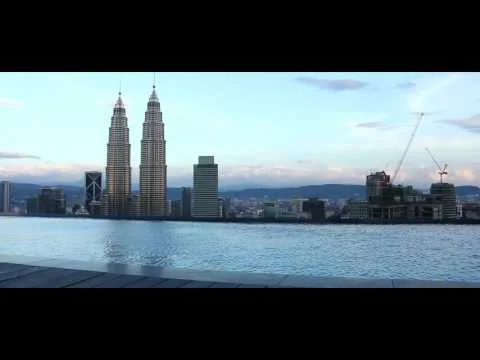 Exterior type: swimming pool deck; features: infinity edge pool, city skyline view, surrounded by urban landscape, visible high-rise buildings, Petronas Twin Towers in view, construction cranes in the background; style: modern.