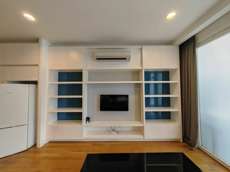 Living features: white built-in shelving unit, wall-mounted TV, air conditioning unit, light wood flooring, large window with sheer curtains, grey curtains, black coffee table, blue accent wall behind shelves; style: modern.