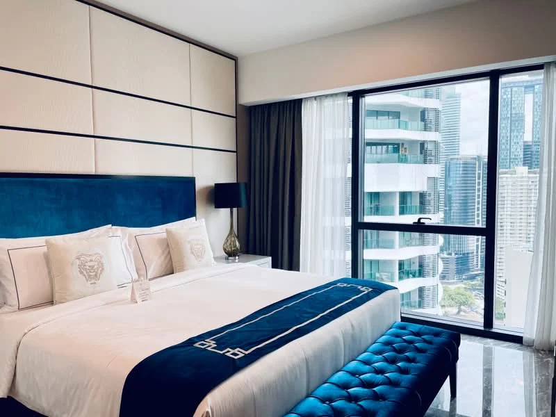 Bedroom features: king-size bed with upholstered headboard, white bedding with blue accents, decorative pillows, nightstand with lamp, blue upholstered bench at foot of bed, floor-to-ceiling windows, curtains, marble flooring, wall paneling with geometric design, city view from window; style: modern; view: city.