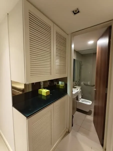 Room features: built-in cabinetry with louvered doors, black countertop, neutral wall colour, tile flooring, recessed ceiling lights, wooden door, bathroom visible in background, white toilet, glass shower enclosure; style: modern.