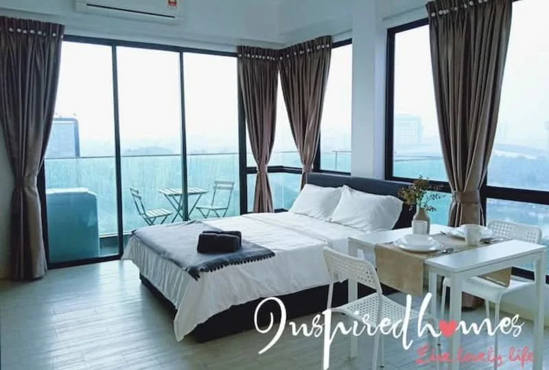 Bedroom features: king-size bed with white bedding, black upholstered bed frame, floor-to-ceiling windows, sheer curtains, wooden flooring, small dining table with two chairs, outdoor balcony with glass railing, city view from balcony, air conditioning unit on wall, decorative throw blanket, bedside table, wall-mounted artwork; style: modern; view: city.