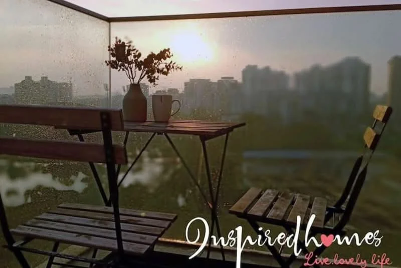 Balcony features: outdoor table, two wooden chairs, potted plant, coffee cup, glass railing, city skyline view, wet surface from rain, sunset lighting; style: modern; view: city.