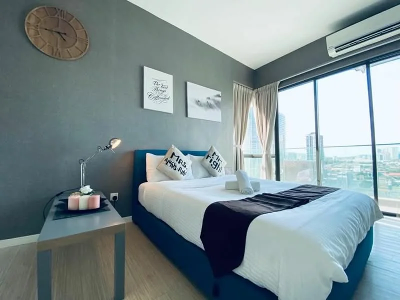 Bedroom features: queen-size bed with white bedding, upholstered headboard in blue, decorative pillows with text, nightstand with grey surface, table lamp, wall clock, framed artwork on walls, floor-to-ceiling windows, sheer curtains, wooden flooring, air conditioning unit; style: modern; view: city.