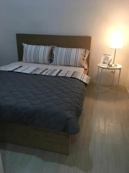 Bedroom features: queen-size bed with striped bedding, wooden bed frame, nightstand with white finish, table lamp with warm light, decorative photo frame on nightstand, light-colored wooden flooring, plain white walls, quilted grey bedspread; style: modern.