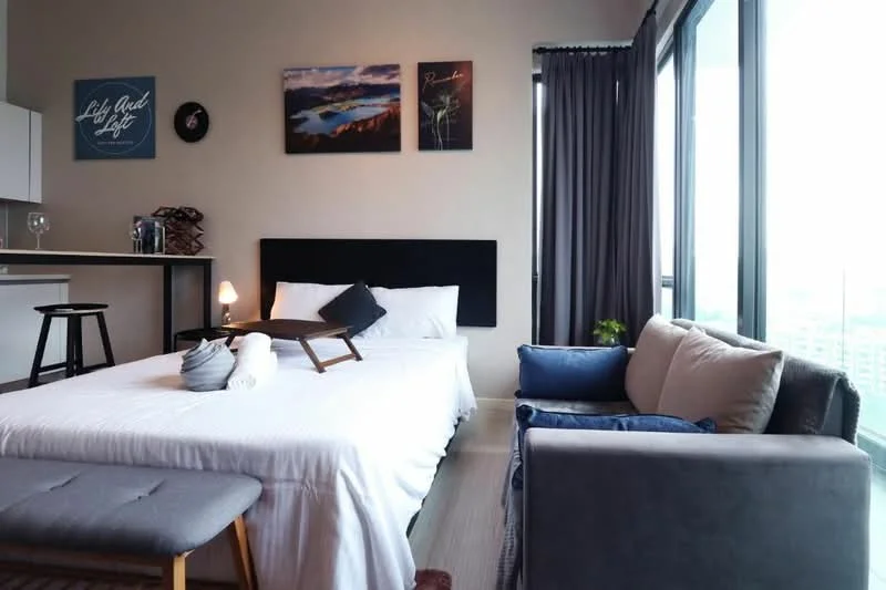 Bedroom features: queen-size bed with white bedding, upholstered headboard, bedside lamp, framed artwork on walls, floor-to-ceiling windows, grey curtains, sofa in grey fabric, throw pillows on sofa, small coffee table, wooden bar stool, open shelving unit, light wood flooring; style: modern; view: city.