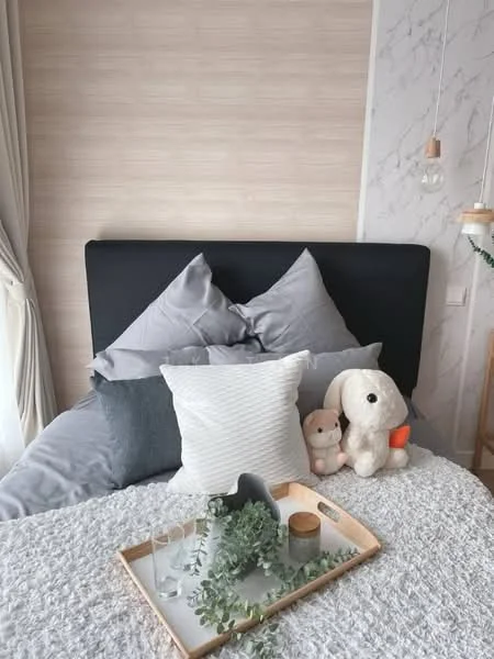 Bedroom features: king-size bed with grey bedding, upholstered headboard, decorative throw pillows in various colors, wooden tray on bed with glasses and a candle, plush toys including a bunny and a bear, light-colored textured wall behind the bed, hanging pendant lights, curtains in soft fabric, wooden bedside table; style: modern.