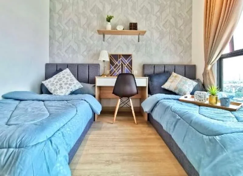 Bedroom features: two single beds with blue bedding, upholstered headboards in grey, wooden desk with decorative items, black chair, shelf above the desk, geometric patterned wallpaper, curtains in light beige, wooden flooring, decorative plants, table lamp; style: modern; view: city.