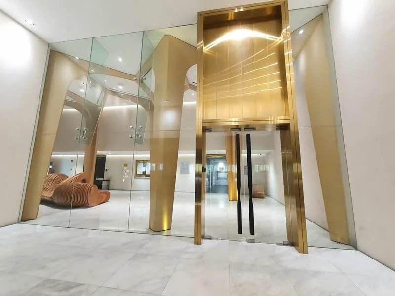Exterior type: main entrance gate; features: glass facade, golden entrance doors, marble flooring, modern architectural design, recessed lighting, wooden seating area, high ceilings, lobby area; style: modern.