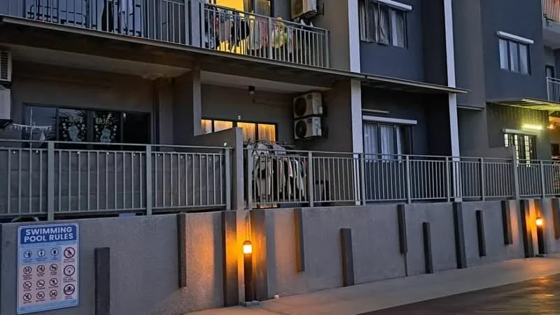Exterior type: low-rise apartment block; features: grey painted facade, balconies with railings, air conditioning units on facade, outdoor lighting fixtures, concrete walkway, swimming pool rules sign; style: modern.