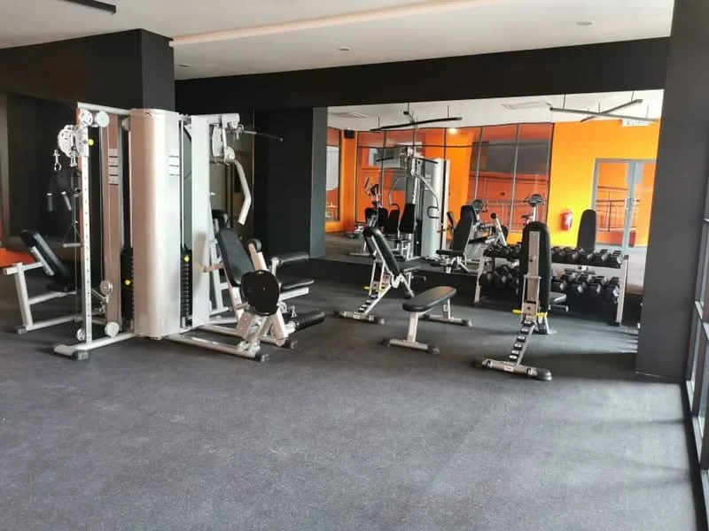 Room features: gym equipment, weight bench, dumbbell rack, multi-station gym machine, black rubber flooring, mirrored wall, orange accent wall, ceiling lights; style: modern.