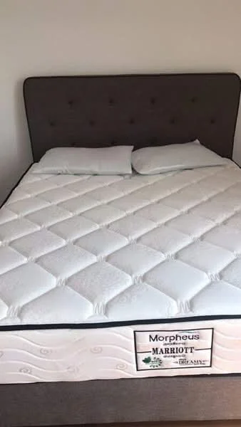 Bedroom features: queen-size bed, upholstered headboard in grey, white bedding, two white pillows, mattress with label, neutral wall color, wooden flooring; style: modern.