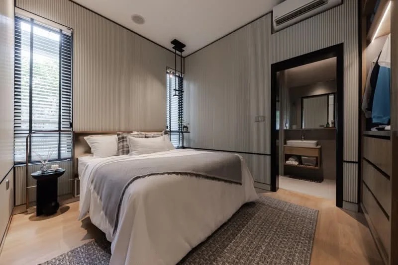 Bedroom features: queen-size bed with white bedding, wooden bed frame, nightstand with glass top, table lamp, two windows with black horizontal blinds, light grey accent wall with vertical paneling, wooden flooring, recessed ceiling lights, wall-mounted air conditioning unit, built-in wardrobe with sliding doors, mirror above vanity, vanity with sink; style: modern.