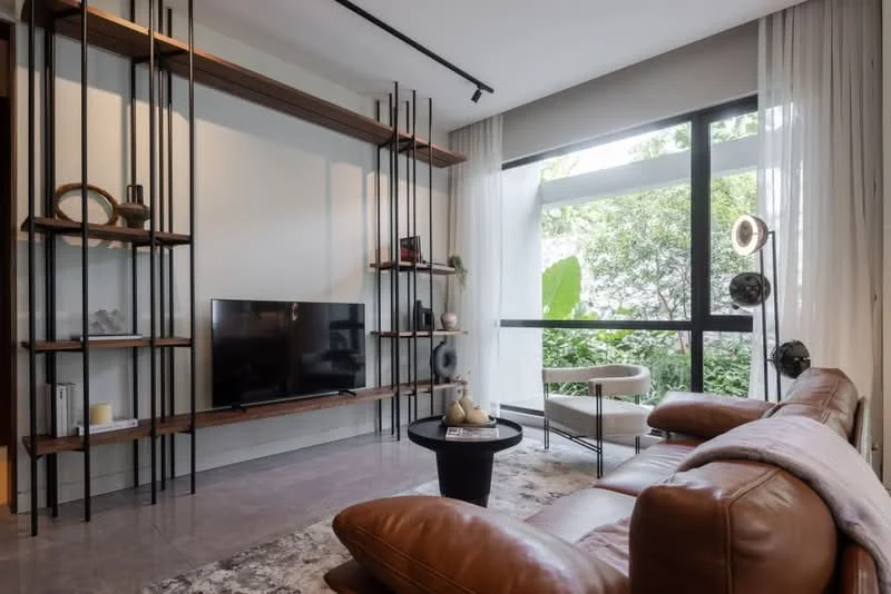 Living features: brown leather sectional sofa, black coffee table, wall-mounted TV, open shelving unit with wooden shelves, decorative items on shelves, large floor-to-ceiling window, sheer white curtains, area rug with geometric pattern, greenery visible outside the window, recessed lighting, track lighting; style: modern; view: garden.