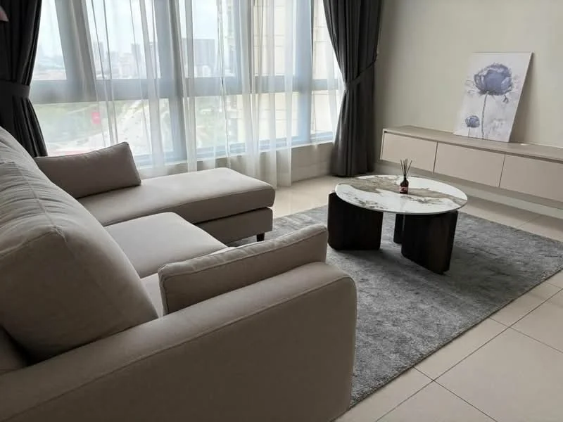 Living features: L-shape sectional sofa in light beige fabric, round coffee table with marble top and dark wooden base, area rug in grey, floor-to-ceiling windows with sheer curtains, light grey walls, TV console in light wood, decorative artwork on wall, natural light from windows; style: modern; view: city.