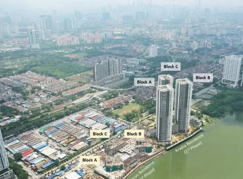 Exterior type: high-rise condominium; features: multiple high-rise buildings, waterfront view, surrounding greenery, construction site visible, urban skyline in background, labelled blocks A, B, C, proximity to other residential areas; style: modern.