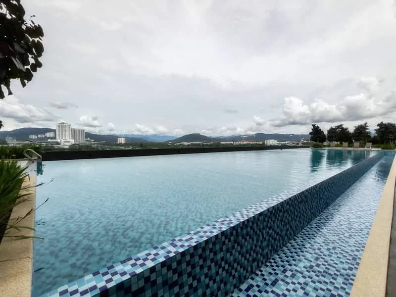 Exterior type: swimming pool deck; features: infinity pool, tiled pool edge with blue mosaic tiles, surrounded by landscaped greenery, view of city skyline, mountain view in background, pool deck with stone paving; style: modern.