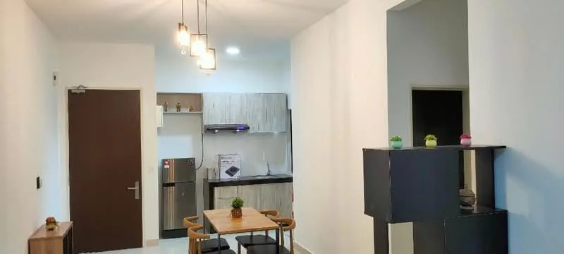 Dining features: 4-seater dining table, wooden dining chairs, pendant lighting with multiple bulbs, white walls, tile flooring, kitchenette with grey cabinetry, stainless-steel fridge, built-in kitchen appliances, open layout, decorative plants on table; style: modern. Room features: door leading to hallway, wall-mounted light fixture, open space leading to kitchen.