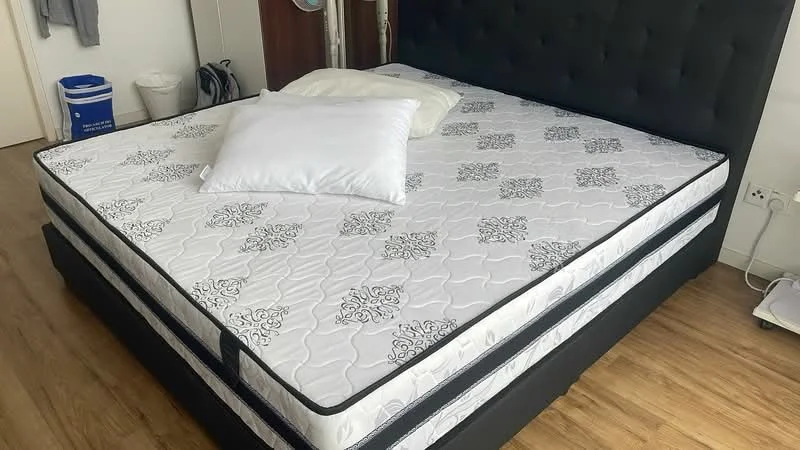 Bedroom features: king-size bed, black upholstered headboard, white mattress with decorative pattern, two white pillows, wooden flooring, bed frame with storage, nightstand with drawer, wall-mounted electrical outlet; style: modern.