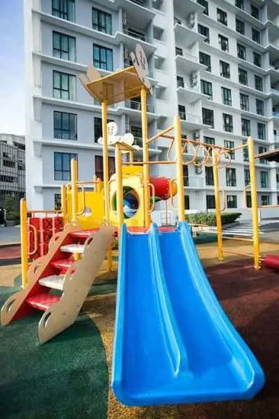 Exterior type: high-rise condominium; features: children's playground, yellow and red play structure, blue slide, climbing frame, rubberized flooring, surrounding landscaping, multiple storey building in background, large windows on building facade; style: modern.